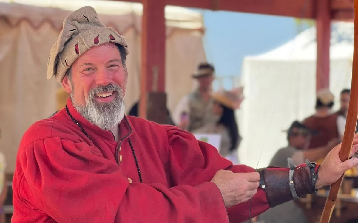 A bearded reenactor in a red medieval tunic drawing a longbow