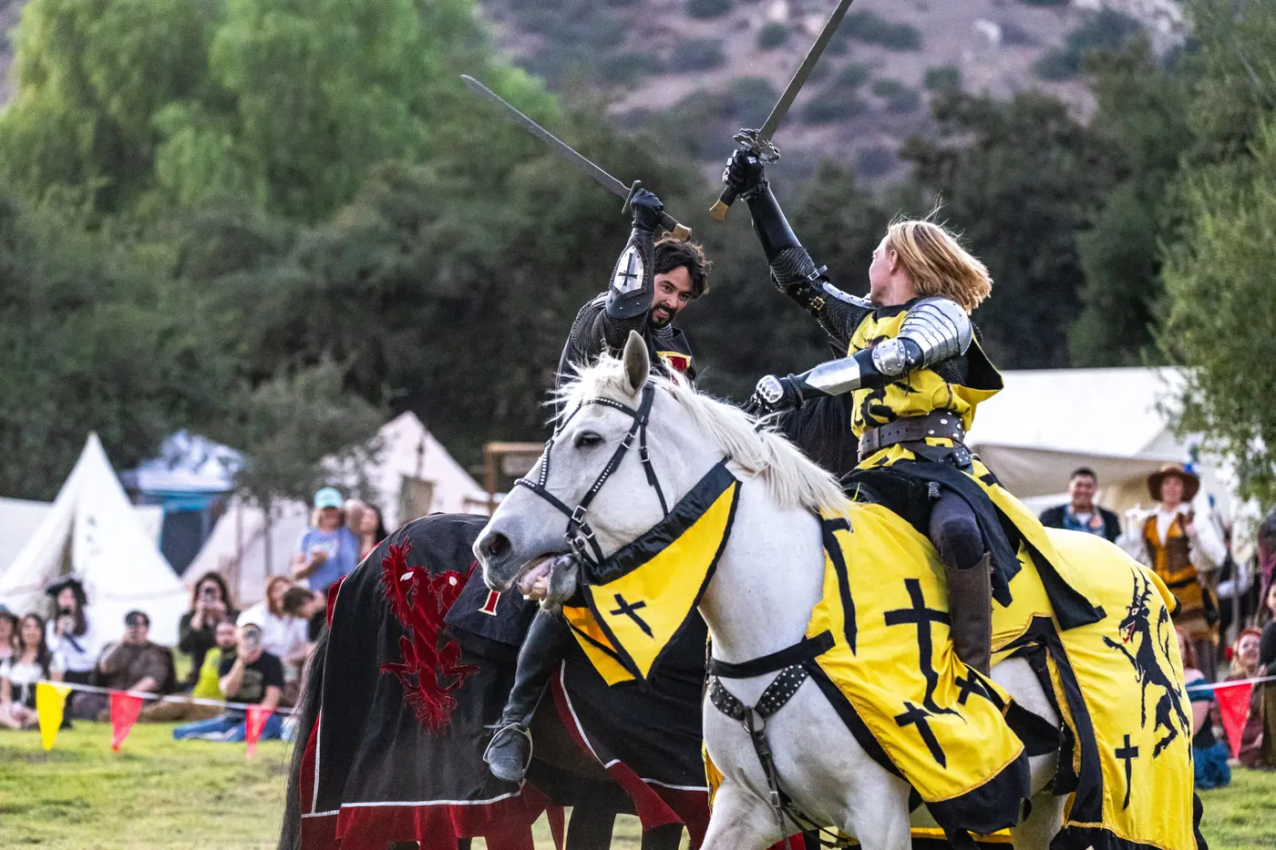 Two armored knights clash at the joust, one horse in yellow and black caparison, as spectators watch from the lists