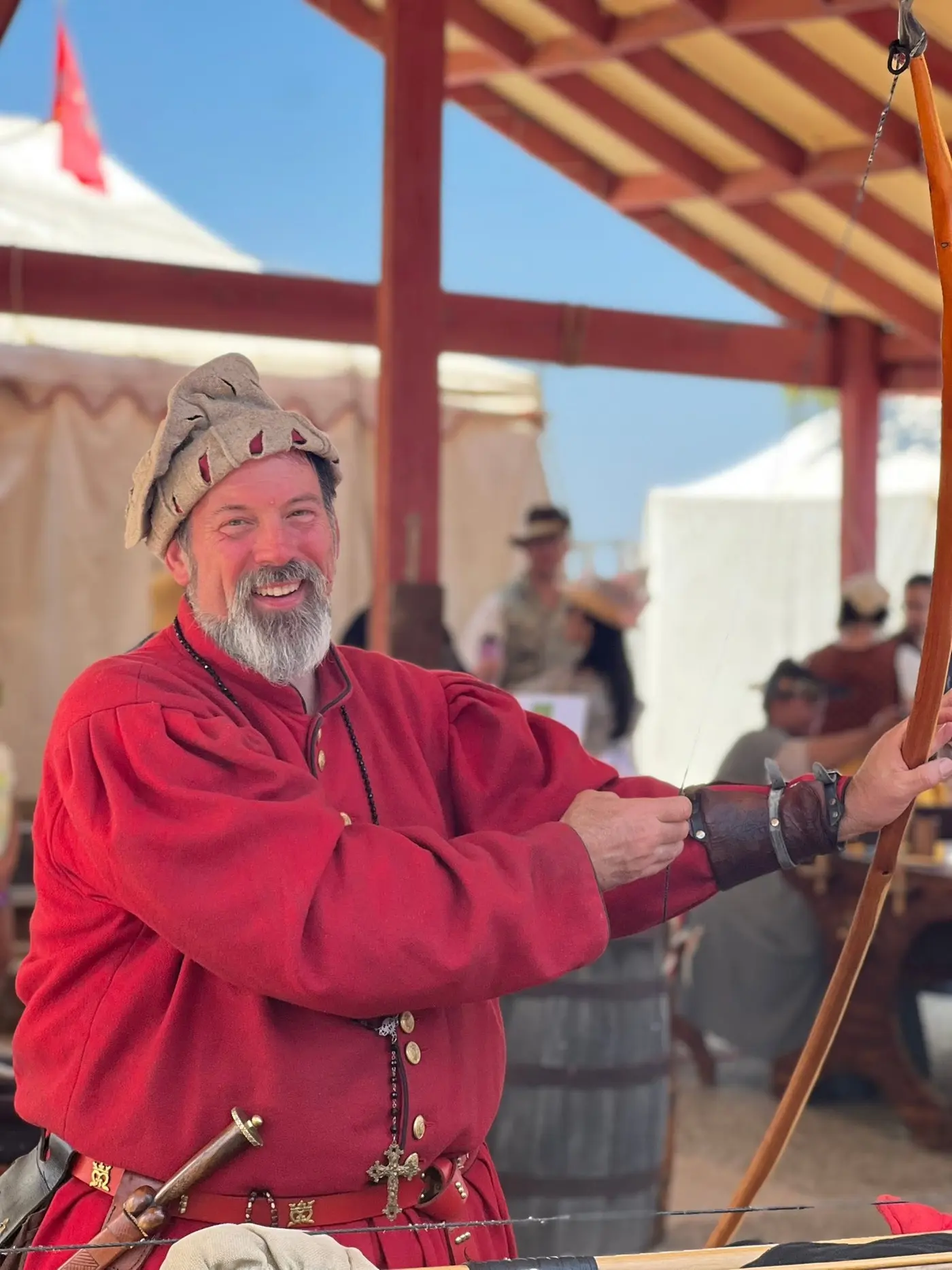 A bearded archer in a red tunic and coif smiles as he draws a longbow