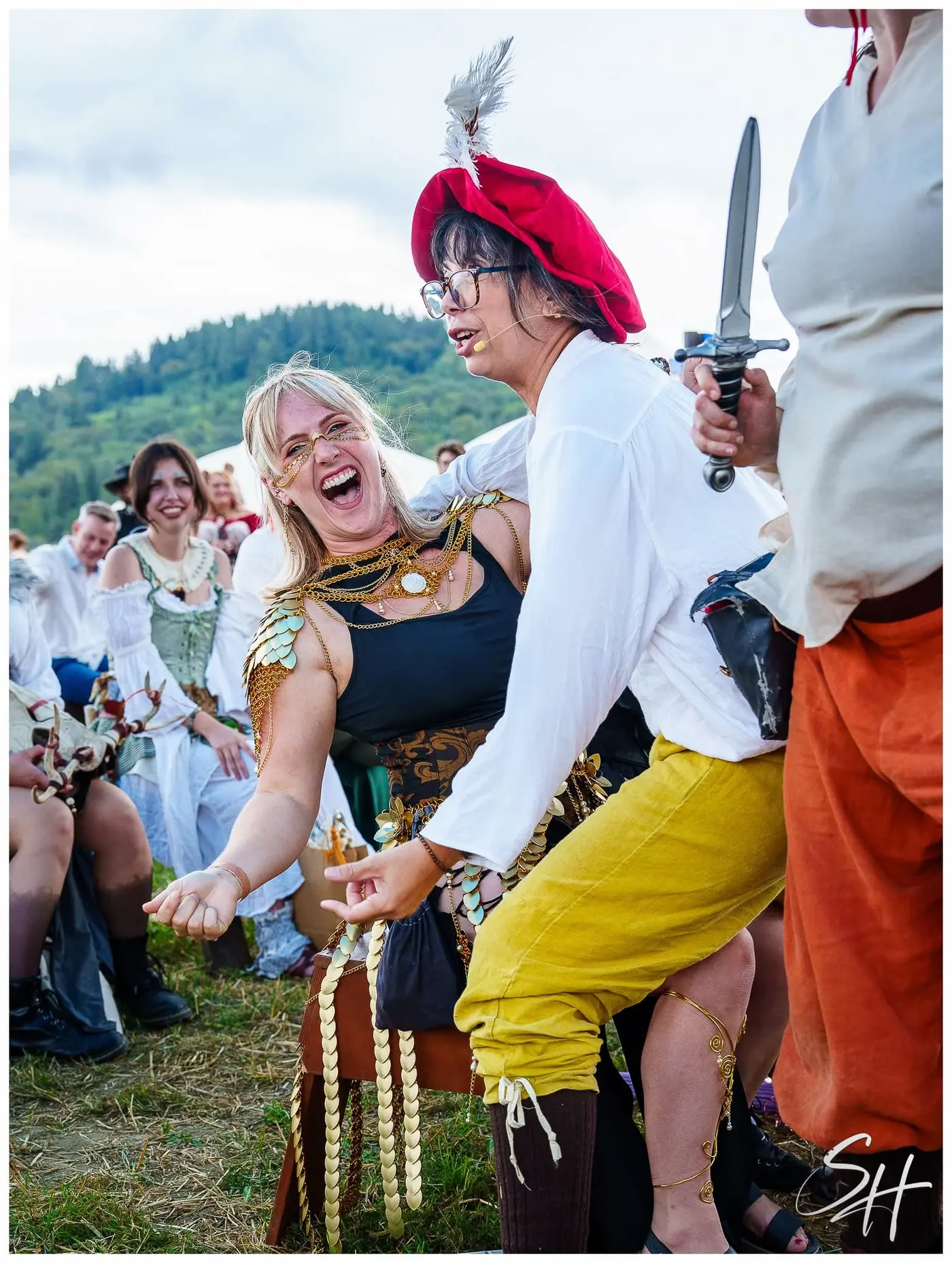 A laughing guest is playfully held at dagger point by a performer in a red cap and yellow hose