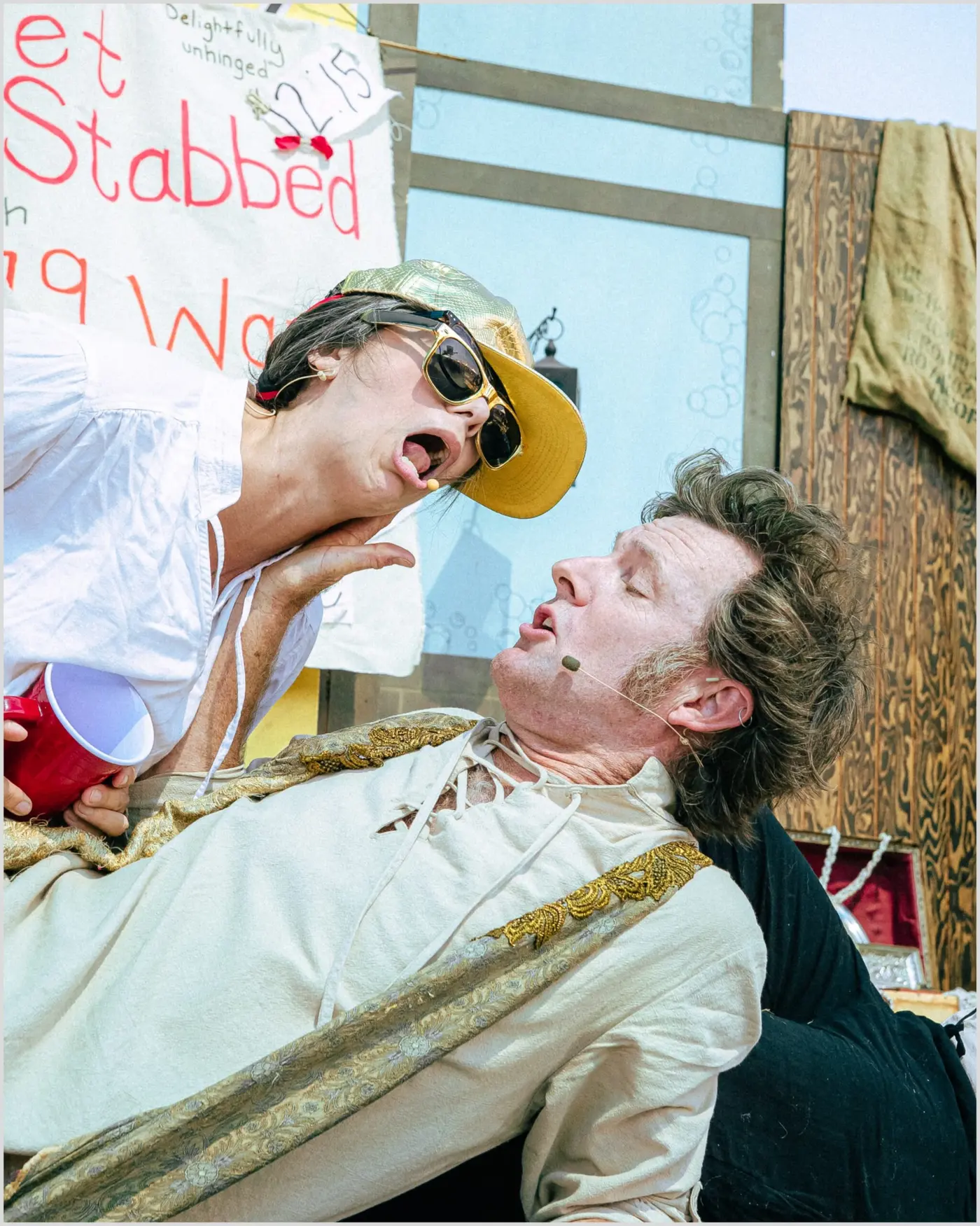 A performer in sunglasses and a cap mock-stabs an actor during a comedy stage show