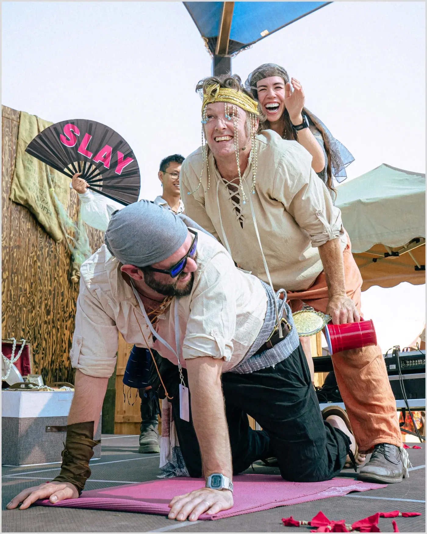 Performers clown on all fours on stage while an actor waves a fan reading SLAY