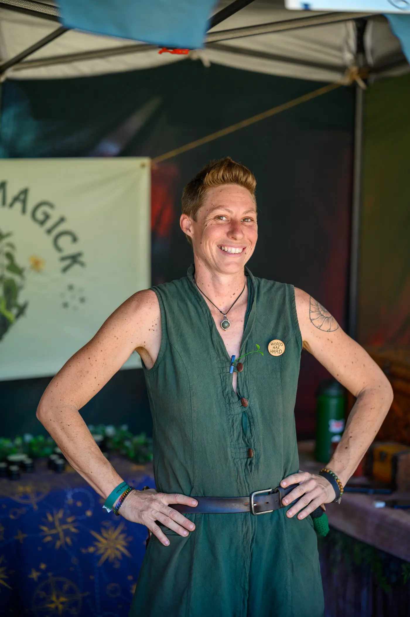 A smiling merchant in a green sleeveless tunic at her Magick booth