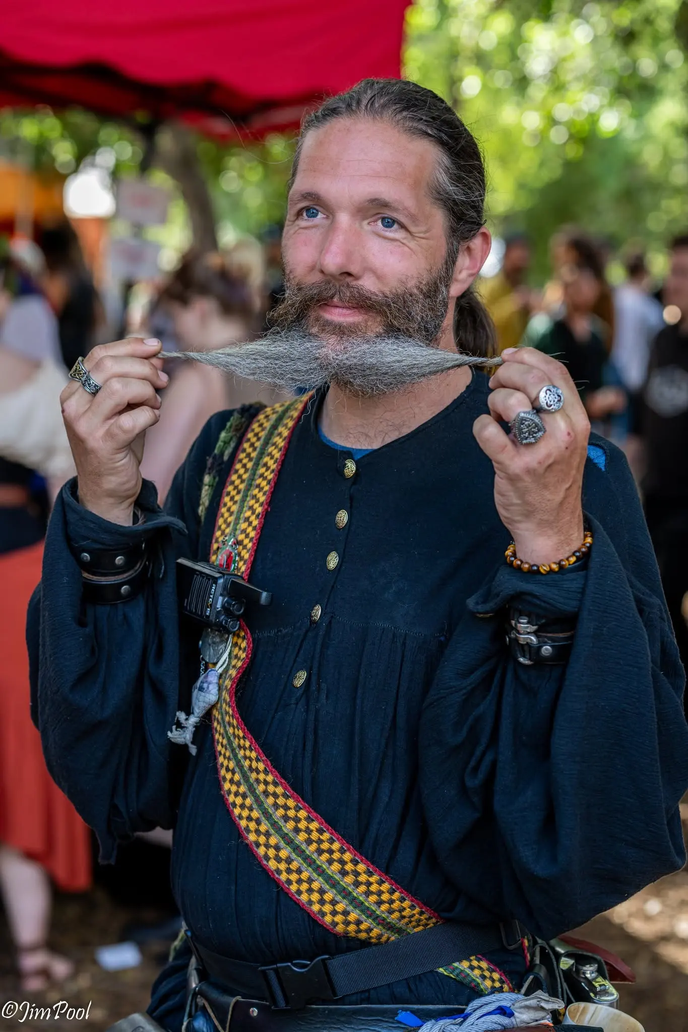 A man in a dark tunic and woven baldric twirls his long mustache