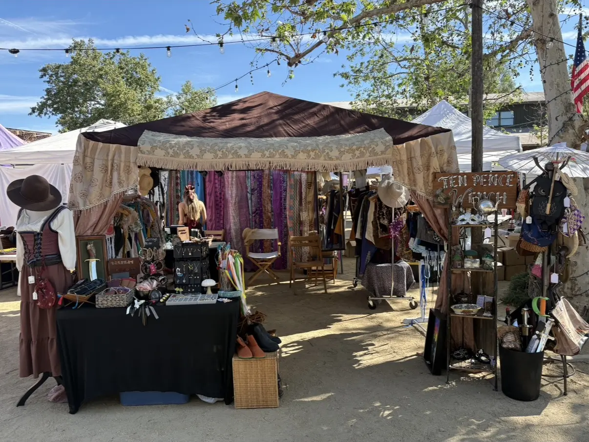 A draped merchant pavilion with fabrics and goods, framed by trees and string lights