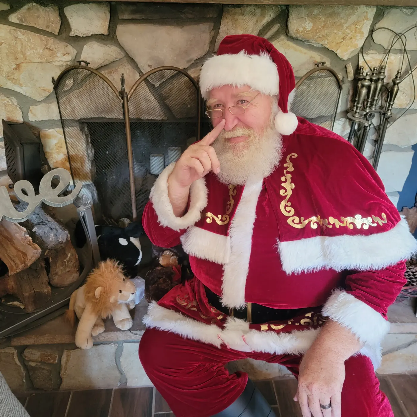 Father Christmas in a red velvet suit and cape, laying a finger beside his nose by a stone fireplace
