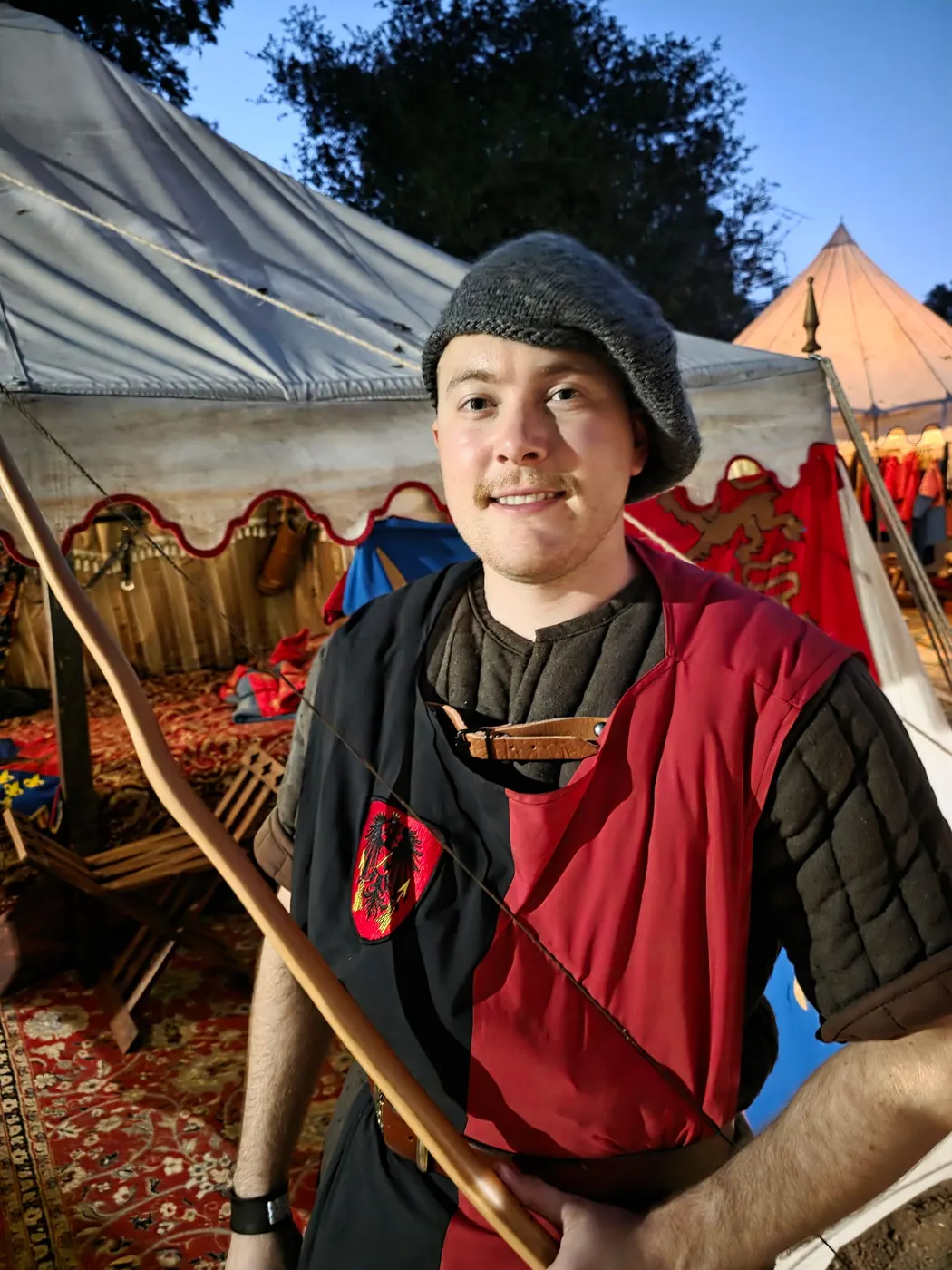 A young archer in a red and black tunic and grey cap holds his longbow before the camp tents