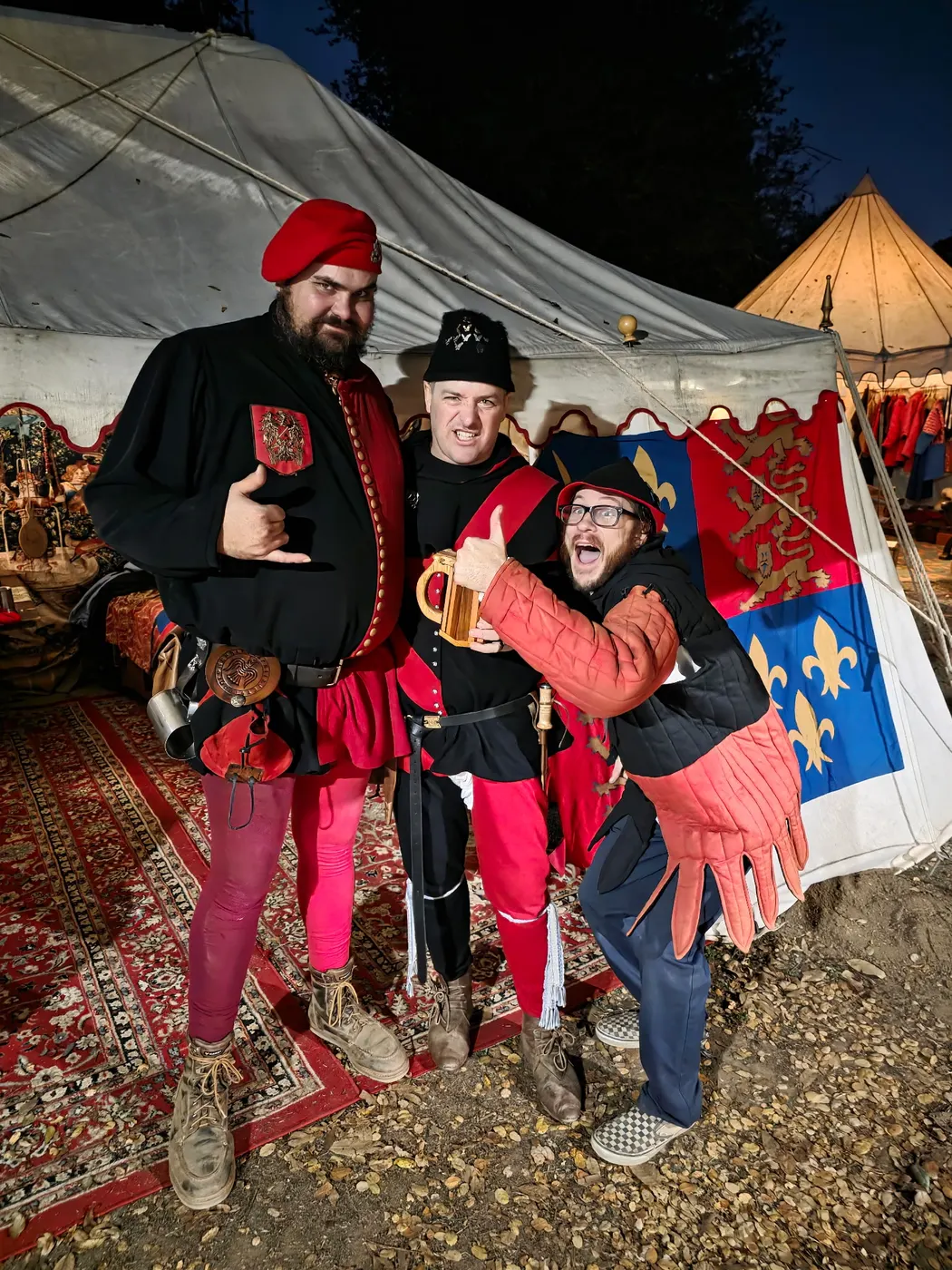 Three reenactors in red and black livery clown for the camera with a tankard before their pavilion and fleur-de-lis banner