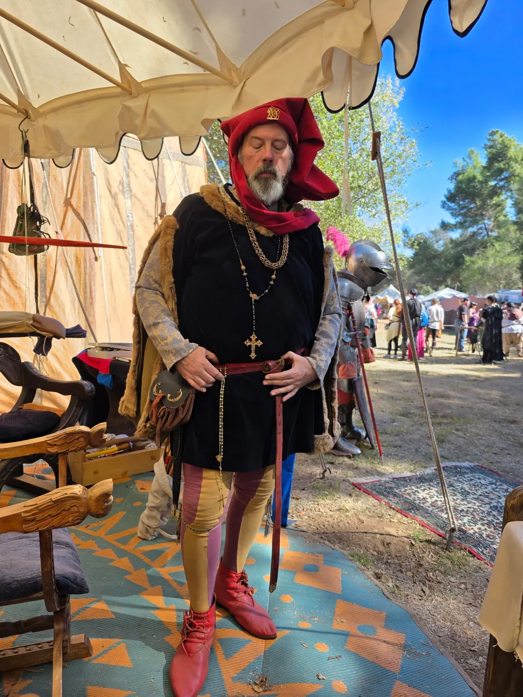 A reenactor in a red chaperon and black doublet with a gold chain and cross on the festival grounds